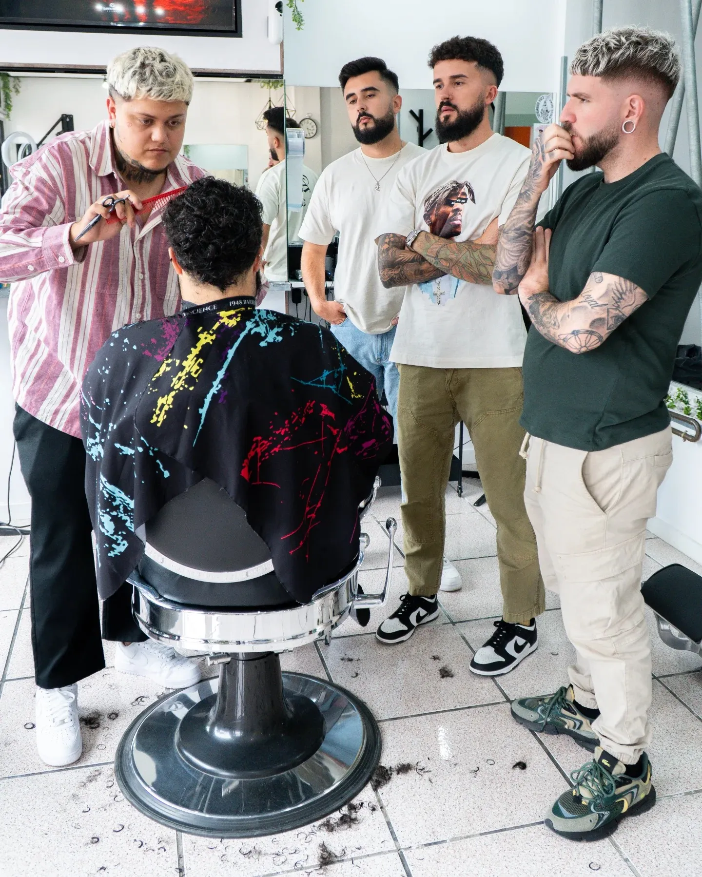 Barbería moderna con cuatro hombres, uno en la silla recibiendo un corte de cabello, mientras los otros tres observan. El barbero, con cabello rubio y camisa de rayas, utiliza tijeras. El ambiente es luminoso y contemporáneo, con espejos y decoraciones urbanas.