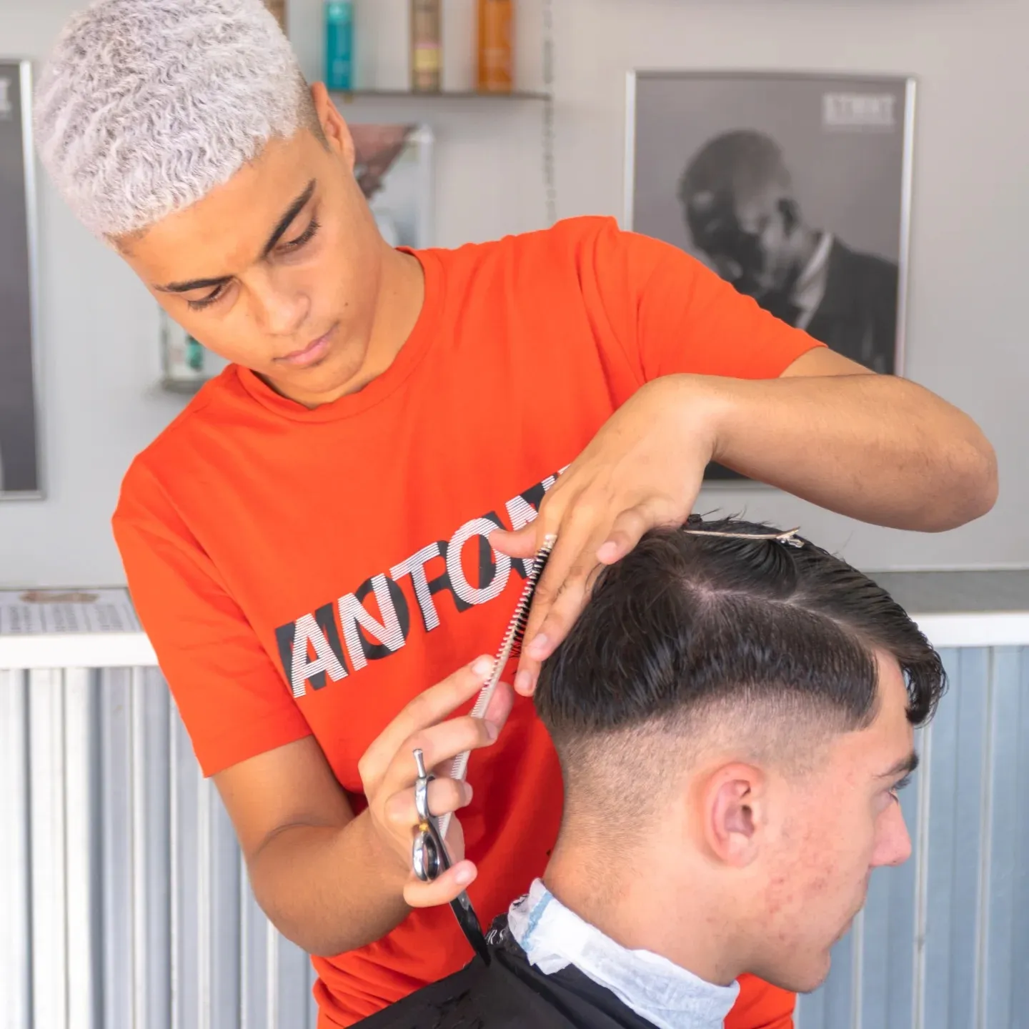 Joven estilista con cabello rizado y rubio corta el cabello de un cliente en una barbería moderna, mostrando técnicas de corte y atención al detalle.